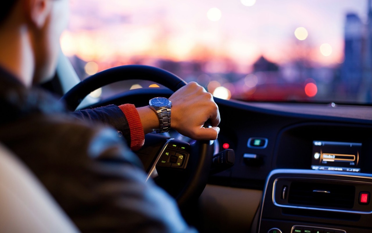 A driver seen from behind, one hand on the wheel, with city traffic ahead at dusk