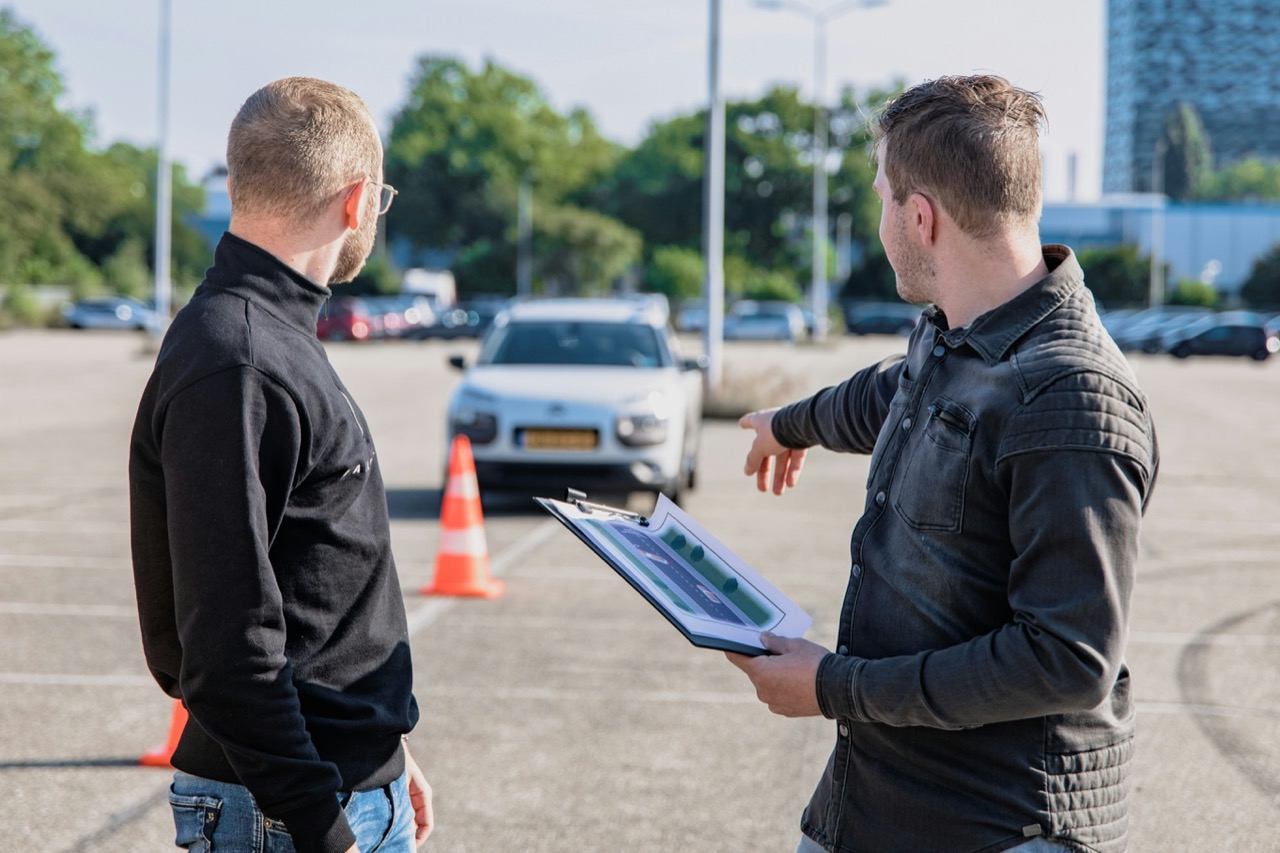 A driving instructor holding a clipboard beside a learner car and traffic cone